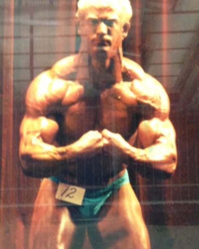 Damian Churchill competing on a bodybuilding stage, wearing competitor number 12